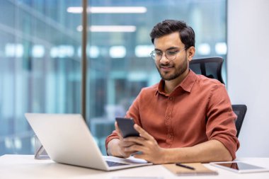 Modern ofiste masasında çalışan gözlüklü genç bir profesyonel akıllı telefon ve dizüstü bilgisayar kullanarak üretkenlik, teknoloji ve iletişim sergiliyor. Resim, rahat bir çalışma ortamı çizer