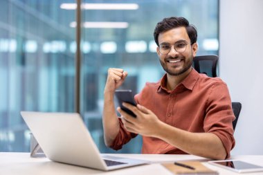 Genç iş adamı ofiste başarıyı kutluyor, akıllı telefon tutuyor, heyecan içinde yumruğunu kaldırıyor. Gülümseyen ofis çalışanları olumlu duygular ve mesleki başarılar sergiliyor.