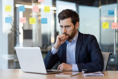 Modern ofiste dizüstü bilgisayarla çalışan takım elbiseli bir iş adamı. İnsan düşünceli, düşünceli ya da karar veren biri gibi görünüyor. Arkaplanda cam duvardaki renkli yapışkan notlar var.