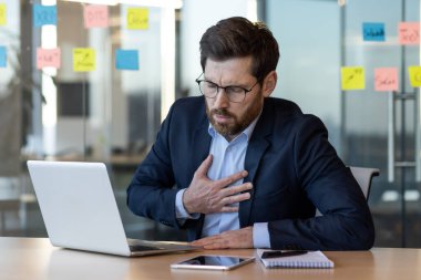 Ofiste dizüstü bilgisayarla çalışırken göğüs ağrısı çeken takım elbiseli bir iş adamı. İş yerinde olası bir sağlık sorunu yaşayan bir ofis çalışanı. Kurumsal çevrede stres ve sağlık sorunları.