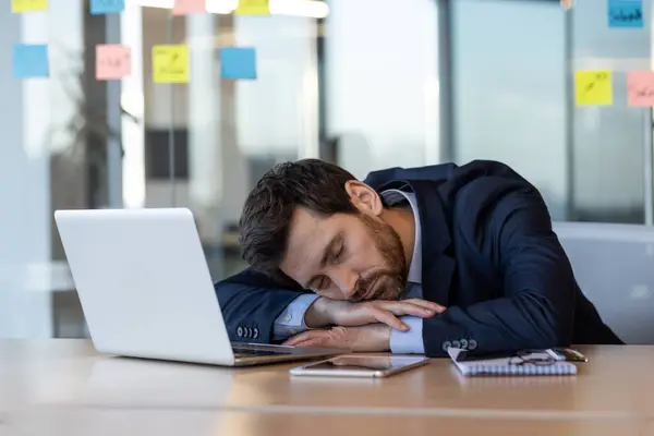 stock image Exhausted businessman in suit sleeping on desk beside laptop and documents in modern office. Concept of workplace fatigue, overwork, stress, and work-life balance among office workers.