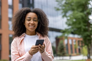 Modern binalar ve arka planda yeşillik olan şehir ortamında akıllı telefonu elinde tutan gülümseyen bir kadın. Günlük kıyafet ve ifade, şehir ortamında rahatlama ve bağlantı olduğunu gösteriyor..