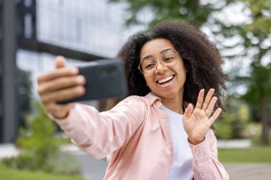 Mutlu genç bir kadın dışarıda el sallarken akıllı telefonuyla selfie çekiyor. Kıvırcık saçlı, gülen kız dışarıda eğleniyor. Eğlence, teknoloji ve sosyal medya kavramı.