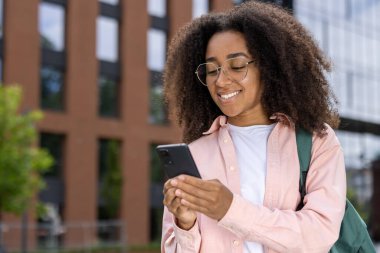 Gözlüklü ve sırt çantalı Afro-Amerikalı kız öğrenci şehir ortamında akıllı telefon kullanırken gülümsüyor. Kendine güvenen, bağlı ve şehir ortamında meşgul..