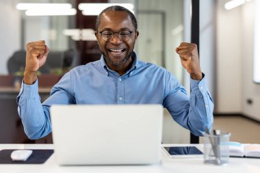 Ofis içi işyerindeki başarıyı kutlayan Afro-Amerikan ofis çalışanı. Olgun, profesyonel bir adam, gözlük takıyor, gülümsüyor ve başarıyla yumruklarını kaldırıyor..