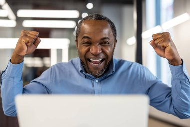 Neşeli Afro-Amerikan ofis çalışanı modern ofis içindeki iş yerinde başarıyı kutluyor. Olgun iş adamı dizüstü bilgisayarla çalışırken yumruklarıyla heyecan ve mutluluk gösteriyor..