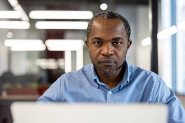 Olgun Afro-Amerikan ofis çalışanı modern ofis ortamında masasında çalışırken odaklanmış görünüyor. Mavi gömlekli profesyonel kıyafet iş ortamını güçlendiriyor..