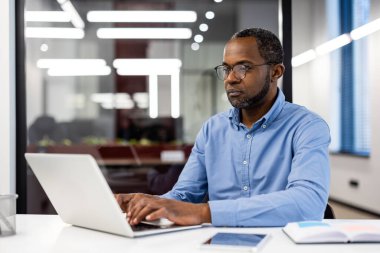 Olgun Afro-Amerikan iş adamı mavi gömlek giyiyor, modern ofisin içindeki iş yerinde laptop kullanıyor. Profesyonel ofis çalışanı iyi aydınlatılmış ofis ortamında çalışmaya odaklanıyor..