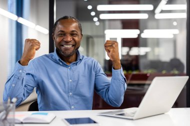 Mutlu Afro-Amerikan ofis çalışanları iş yerinde başarıyı kutluyor. Modern ofiste yumruklarını sıkarak gülümseyen olgun bir iş adamı. Başarı, mutluluk ve profesyonel başarı kavramı.