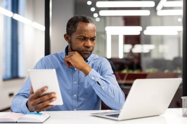 Olgun bir Afro-Amerikan ofis çalışanı. Tablet ve dizüstü bilgisayardaki verileri analiz ediyor. İşadamı modern ofis içinde çalışmaya odaklanmış, konsantrasyon ve verimliliği gösteriyor.