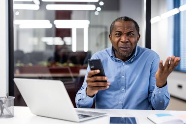 Olgun Afro-Amerikan ofis çalışanı iş yerinde akıllı telefon kullanırken hayal kırıklığına uğradı. Ofis masasında profesyonel kıyafetli bir iş adamı kafası karışmış görünüyor. İş stresi kavramı