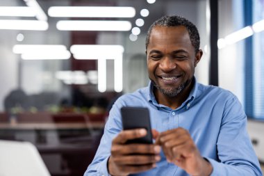 Olgun Afro-Amerikan iş adamı modern ofiste akıllı telefon kullanıyor. Gülümseyen ofis çalışanı iş günü cep telefonuyla meşgul oluyor. Profesyonel işyeri ortamı