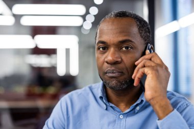Olgun bir Afrikalı Amerikalı ofis çalışanı modern ofisin içinde telefonla konuşuyor. Profesyonel iş adamı telefon görüşmesi yapıyor, odaklanmış ifade.