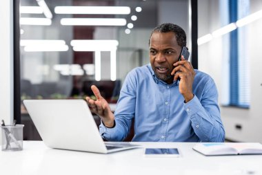 Olgun iş adamı, modern ofiste telefonda önemli konuları tartışıyor. Ofis çalışanı masadaki dizüstü bilgisayar, defter ve tablete kızgın ya da üzgün görünüyor. Profesyonel işyeri ortamı.