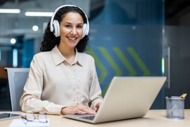 Modern ofiste dizüstü bilgisayar kullanan kulaklık takan mutlu bir kadın. Uzaktan kumandalı işçi çalışırken online toplantı, video görüşmesi ya da müzik yapmaktan hoşlanıyor. Şık dekorlu, parlak, profesyonel çalışma alanı.