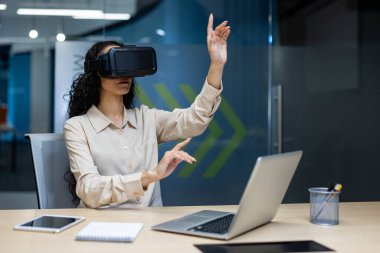 Modern ofiste sanal gerçeklik kulaklığı kullanan bir iş kadını, dijital arayüzle etkileşimde. VR teknolojisine dalmış genç bir kadın, sanal çalışma alanını keşfediyor, iş dünyasında yenilikler yapıyor.