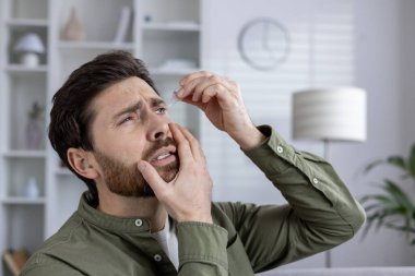 Man applying eye drops for dry and irritated eyes relief indoors. Person wearing green shirt using medication for eye discomfort in natural environment. Health, wellness, and self-care concept. clipart