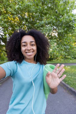 Yoga minderli gülümseyen kadın dışarıda yürürken telefonla konuşuyor. Temiz havanın ve doğanın tadını çıkarıyor. Günlük spor kıyafetleri ve kulaklıklar takıyor..