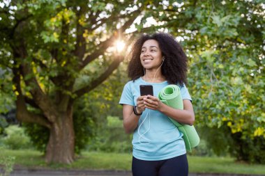 Genç Afrikalı Amerikalı atlet yoga minderi taşıyor, müzik dinliyor, dışarıda parkta akıllı telefon kullanırken gülümsüyor. Spor meraklısı doğa, teknoloji ve sağlıklı yaşam tarzını seviyor..