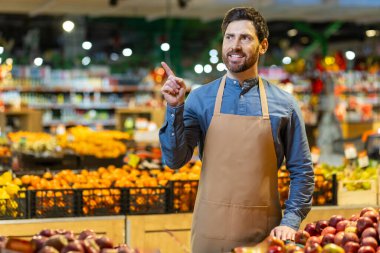 Gülümseyen market çalışanı meyve reyonunda dikilip bir şeyi işaret ediyor. Çalışan önlük takıyor, müşterilere ya da meslektaşlara bir şey gösteriyor, taze meyve ve sebzelerle çevrili.