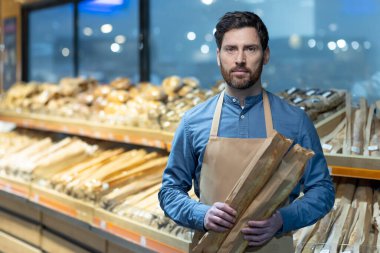 Baker marketin ekmek reyonunun önünde taze baget tutuyor. Önlük giyip, arka planda pişmiş mallarla fırın koridorunda dikilmek. Meslek, ekmek, fırın, market konsepti.