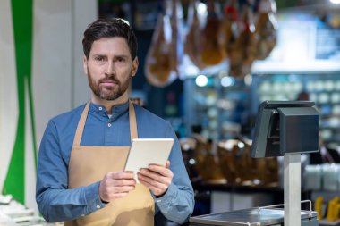 Marketteki kasada dijital tablet kullanan bir mağaza çalışanı. Satışları veya envanteri yönetmek için elektronik alet kullanan bir adam. İçerde, odaklanmış ifade, perakende çalışma ortamı.