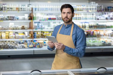 Erkek süpermarket çalışanı markette dijital tablet kullanarak envanteri kontrol ediyor. Çalışan çeşitli ürünlerle rafların önünde duruyor, envanter yönetimine odaklanmış..
