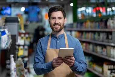 Gülümseyen bir süpermarket çalışanı mağaza koridorundaki envanter yönetimi için dijital tablet kullanıyor. Çalışanlar önlük giyer, tablet taşır, envanter yönetimi görevlerini yerine getirirler. Modern perakende teknolojisi