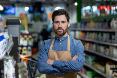 Kendine güvenen erkek mağaza çalışanı bir süpermarket koridorunda kolları bağlı duruyor. Önlük giyen işçi kameraya ciddi bir ifadeyle bakıyor. Arkaplanda ürünlerle dolu raflar.