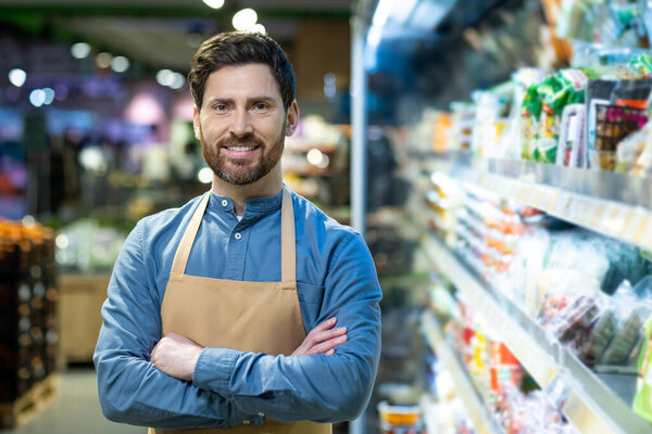 Confident male grocery store employee standing with arms crossed in supermarket aisle. Worker wears apron and smiles at camera surrounded by various goods. Professionalism and retail work concept.