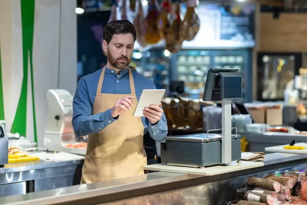Market çalışanı kasada tablet kullanıyor. Önlük giyen işçiler, modern süpermarkette durup envanter ve cihazların fiyatlarını kontrol ediyorlar..