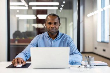 Modern ofiste dizüstü bilgisayarla çalışan kendine güvenen bir iş adamı. Bilgisayar ve fare kullanarak profesyonelce odaklanmış, çalışma görevleri yapıyor. Parlak, modern ofis geçmişi.