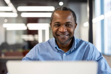 Modern ofis ortamında dizüstü bilgisayarda çalışan gülümseyen bir iş adamı. Profesyonel görünüş, üretken görevler üstlenmek, olumlu tavır sergilemek. Parlak çalışma alanı odaklanmayı ve verimliliği arttırır