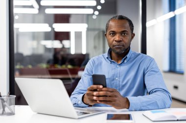 Modern ofiste dizüstü bilgisayarla çalışırken akıllı telefon kullanan odaklanmış bir işadamı. Profesyonelce çoklu görev ve iş iletişimi. Çağdaş bir çalışma alanında işletme yöneticisi.