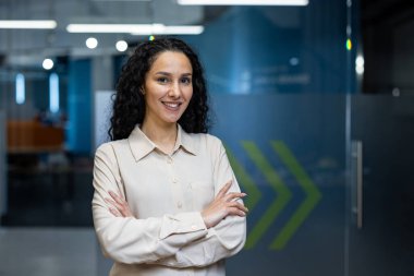 Modern ofiste kollarını kavuşturmuş kıvırcık saçlı kendine güvenen bir iş kadını. Profesyonel görünüş ve olumlu ifade. Liderlik kavramı, başarı ve işyeri ortamı.