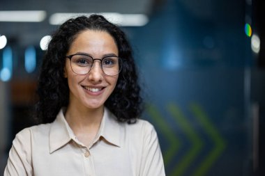 Modern ofis ortamında gülümseyen kendinden emin genç bir iş kadını. Profesyonel ofis çalışanı, gözlük takıyor ve günlük kıyafetler giyiyor. Arka planda mavi ve yeşil renklere sahip modern işyeri kurulumu yer alıyor.