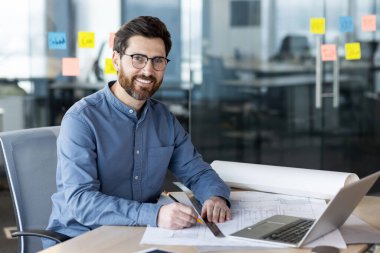 Mavi gömlekli gülümseyen mimar ofis masasında dizüstü bilgisayar ve arka planda yapışkan notlar üzerinde çalışıyor. Planlama, tasarım, inşaat ve modern işyeri ortamı kavramı.