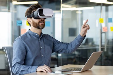 İş adamı ofiste dizüstü bilgisayarda çalışan sanal gerçeklik kulaklığı kullanıyor. VR teknolojisini keşfetmek, yenilikçi etkileşim, işyeri yaratıcılığı. Gündelik giysiler modern çalışma ortamını yansıtıyor