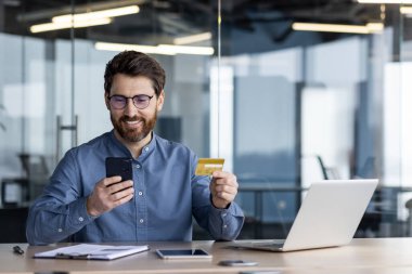 Gülümseyen işadamı elinde kredi kartı tutarken akıllı telefonu kullanıyor. Modern ofis ortamında dizüstü bilgisayar, telefon ve belgelerle birlikte masada oturuyor..