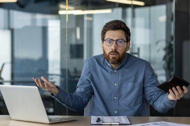 Kafası karışmış işadamı, modern ofiste dizüstü bilgisayarla otururken telefonu tutuyor. Profesyonel ortamda hayal kırıklığı, stres ve kafa karışıklığı