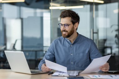 Modern ofiste belgelerle ve bilgisayarla çalışan gülümseyen bir iş adamı. Profesyonel analiz evrakları, veriler ve çizelgeler. Başarılı girişimci planlama stratejisi ve masalarda hedefler