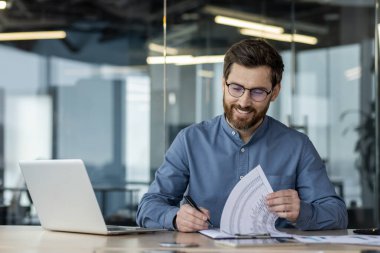 Belgeleri inceleyen ve modern ofis alanında dizüstü bilgisayarla çalışan kendine güvenen bir iş adamı. Üretkenliği ve başarıyı teşvik eden profesyonel ortam.