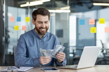 Modern ofis ortamında para sayarken gülümseyen bir iş adamı dizüstü bilgisayar ve arka planda yapışkan notlar ile. Mesleki çevrede başarı, finansal büyüme ve mutluluk kavramı.