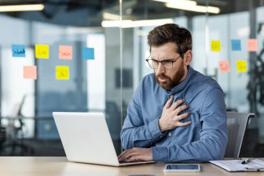 Gözlüklü ve sakallı bir işadamı ofis masasında dizüstü bilgisayarın önünde oturuyor ve sandığı rahatsızlık içinde tutuyor. Stresli çalışma ortamı ve arka plandaki cam duvara yapışmış not kağıtları.