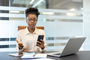 Modern ofisteki genç Afrikalı Amerikalı iş kadını telefon görüşmesinde endişeli görünüyordu. Bilgisayarlı ofis ayarları ve masadaki belgeler. Duygular ve modern iş iletişimi