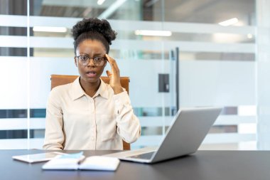 İş yerindeki genç Afrikalı Amerikalı iş kadını kafası karışmış görünüyor. Masasında dizüstü bilgisayarı ve telefonu olan, kafası karışmış gözlüklü bir kadın.