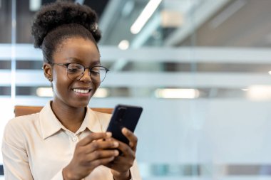 İş yerinde telefonu kullanan ve gülümseyen genç Afrikalı Amerikalı iş kadını. Mutlu şirket çalışanı çevrimiçi iş ortamından keyif alıyor.
