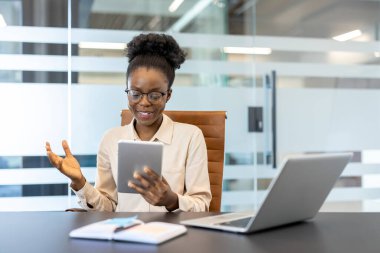 Modern ofisteki genç Afrikalı Amerikalı iş kadını tabletle görüntülü görüşme yapıyor. Profesyonel etkileşim, dijital iletişim ve işyeri ortamı.