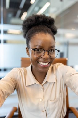 Genç Afrikalı Amerikalı iş kadını ofis ortamında kameraya bakıyor. Profesyonel kıyafet, gülümseyen, mutlu, kendine güvenen, telefonu kullanan. Ofis alanı, modern dekor, doğal ışıklandırma.