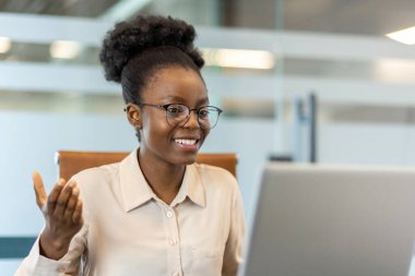 Genç Afrikalı Amerikalı iş kadını iş yerinde video görüşmesi yapıyor. Modern ofis içinde profesyonel iletişim için bilgisayar ve telefon kullanan kendine güvenen bir şirket çalışanı.